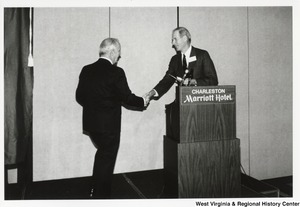 Governor Arch Moore walking onto a stage in the Charleston Marriott Hotel and shaking the hand of an unidentified man who was announcing him to the stage.