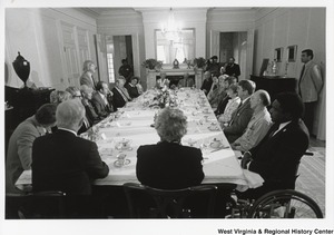 Governor Arch Moore (first on left, with back turned) having a meeting with an unidentified group of people. They are all seated around a large square table and have cups and saucers in front of them. An unidentified man is standing halfway down the row speaking. A camera man is filming the event from a doorway in the right corner.