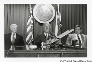 Governor Arch Moore (center) speaking to the press. The Governor is holding what appears to be a piece of wood in his hands. Two unidentified men are sitting on either side of the governor.