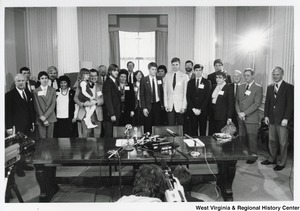 Governor Arch Moore (first on left) with an unidentified group of people posing for the press. They are standing behind a table with press microphones in the center. A cameraman can be seen in the bottom center of the photograph filming the group.