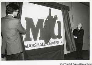Governor Arch Moore (right) and Marshall University President Dale Nitzschke (left) unveiling the new logo for Marshall University. The logo features a capitalized M and U with the outline of the state of West Virginia behind it and the words Marshall University at the bottom.