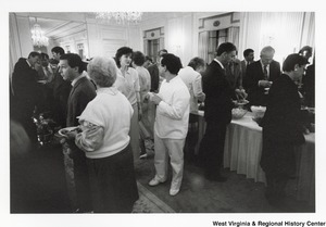 A group of people getting food from a buffet. Other people are mingling while eating.