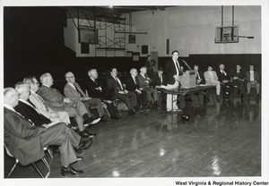 An unidentified man is giving a speech in a gymnasium. Sitting in a curve around the podium is a group of unidentified men. Governor Arch Moore is seated second to the left of the speaker.