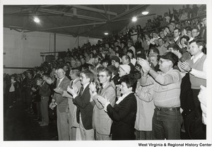 A large crowd of people standing in the bleachers of a gym clapping.