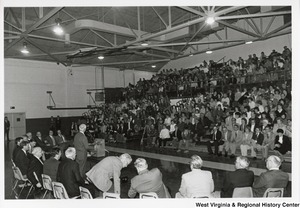 Governor Arch Moore giving a speech in a gymnasium. Sitting in a curve behind him is a group of unidentified men. The photo is taken from behind the men and Governor Moore and the filled bleachers are visible.