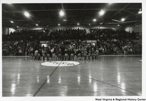 An unidentified man is giving a speech in a gymnasium. Sitting in a curve behind him is a group of unidentified men, including Governor Arch Moore who is sitting directly behind the man. The center of the gymnasium floor has a white circle with WHS in the center. The photo is taken from a distance behind the men and Governor Moore and the filled bleachers are visible.