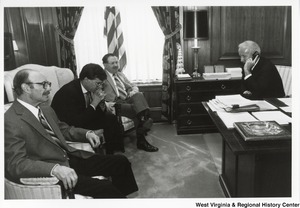 Governor Arch Moore seated at a desk on the telephone. Three unidentified men are seated in front of the Governor.