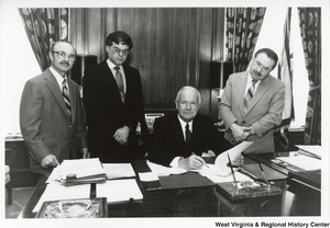 Governor Arch Moore seated at his desk with his pen poised about a document. Three unidentified men are standing behind the desk with the Governor.