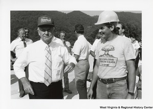 Governor Arch Moore standing with an unidentified man from Demtech Demolition. Governor Moore is wearing a hat that reads Explosive Demtech Demolition. The unidentified man is wearing a hardhat and a shirt that reads: The Explosive Problem Solver 1-717-224-4017. A group of unidentified people are standing behind the Governor mingling.