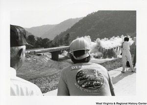 Governor Arch Moore (left corner) standing with an unidentified man from Demtech Demolition watching the demolition of a bridge. The unidentified man is wearing a hard hat and a shirt saying Demtech Wayne Drilling & Blasting.