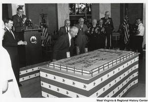 Governor Arch Moore leaning over a large sheet cake for the 350th anniversary of the National Guard. The cake has the Minute Man Statue on the right side and Happy Birthday National Guard 350 Years 1636-1985 written on the rest of the cake. In the background men in uniform are clapping.
