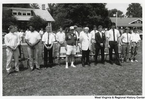 Governor Arch Moore (first row, fifth from the left) standing outside with an unidentified group of men. All the men are standing in front of their seats on the grass. go