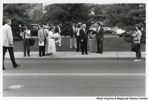 Governor Arch Moore talking to an unidentified man on a sidewalk while being filmed. Multiple people are filming the interaction and a group of people are standing by watching.