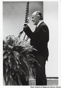 An unidentified man standing at a podium speaking. A large potted plant is on the podium.