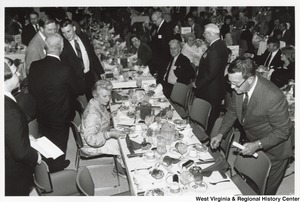 Governor Arch Moore standing behind Shelly and shaking the hand of an unidentified man. Shelley Moore is seated at the table in front of the Governor. There are rows of tables filled with people.