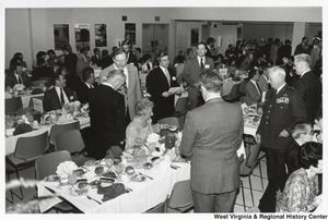 Shelley Moore seated at the first table moving a bowl of food during a dinner. Governor Arch Moore is standing behind Shelley talking to an unidentified man. Surrounding the Governor and his wife are people seated at other tables while others mingle among the tables.