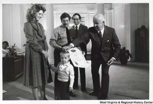 Governor Arch Moore rubbing a little boys head. The boy is holding onto the skirts of an unidentified woman.  Standing beside the Governor is two unidentified men. The man in the middle is holding an official document from the State of West Virginia.