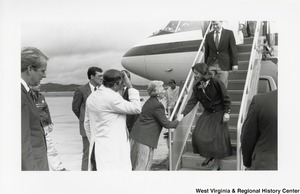 Shelley Moore is shaking the hand of an unidentified woman who is disembarking a plane. Another unidentified man is coming down the stairs behind the woman. An unidentified man is standing with Shelley to greet the guests.