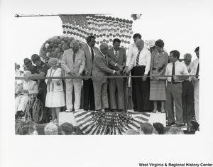 Governor Arch Moore (seventh from the left) cutting a ribbon with Senator Robert C. Byrd (fifth from the right) and two other unidentified men. They are standing on a platform with other unidentified people and a balloon flag is hanging up behind them.