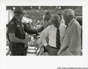 Governor Arch Moore listening to an unidentified man at a large event outside. A state police officer is standing with them.