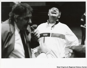 Governor Arch Moore wearing a FILA track suit and laughing. An unidentified man is in front of him and appears to be speaking into a microphone.
