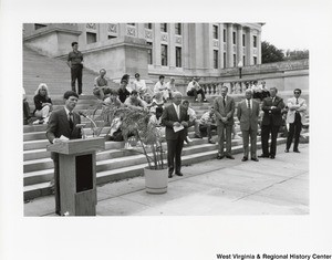 An unidentified man is giving a speech on the steps of a building. Governor Arch Moore is standing to the right of the man with four other men. People are sitting on the steps behind the speaker and the Governor.