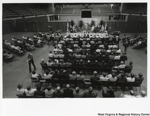 An unidentified man is giving a speech. Governor Arch Moore is standing beside him. The photograph is taken from elevated seating looking down upon the seated crowd and the podium.