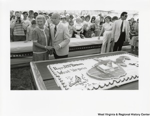 Governor Arch Moore and Shelley Moore standing beside a large sheet cake celebrating the 125th birthday of the state. Behind the Governor is a crowd of people waiting to celebrate and have a piece of cake.  The cake features the state of West Virgnia in the center with the capitol building in the center of the state. On the left corner of the cake is a cardinal and above it is the words Happy 125th Birthday West Virginia.