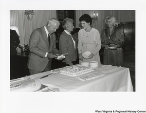 Governor Arch Moore cutting a cake. Shelley Moore is standing beside him talking to two unidentified women.