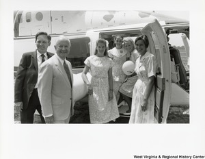 Governor Arch Moore (second from left) standing beside a plane with one man and four women. The four women are standing in the doorway of the plane.