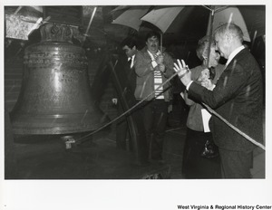 Governor Arch Moore ringing the replica Liberty Bell at the State Capitol. Shelley Moore and two unidentified men are standing with Governor Moore. They are all using umbrellas because its raining.