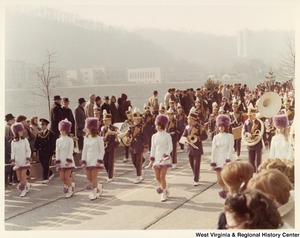 A unidentified high school marching band during Governor Arch Moores inaugural parade in Charleston. The band is wearing either blue or black marching band uniforms with yellow/gold accents. The majorettes in front of the band are wearing white dresses with brown fur accents on the wrist and hem. They are also wearing brown fur hats.