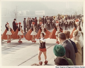 An unidentified high school during Governor Arch Moores inaugural parade in Charleston. The band is wearing black and white uniforms with a red s on the chest. The majorettes are wearing white and black dresses with red capes and white hats with a red plume.